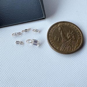 May include: A collection of small, clear, rectangular-cut gemstones with silver-colored settings. The gemstones are scattered on a textured white surface next to a US one-dollar coin. The gemstones vary in size and shape, some with loops for hanging.