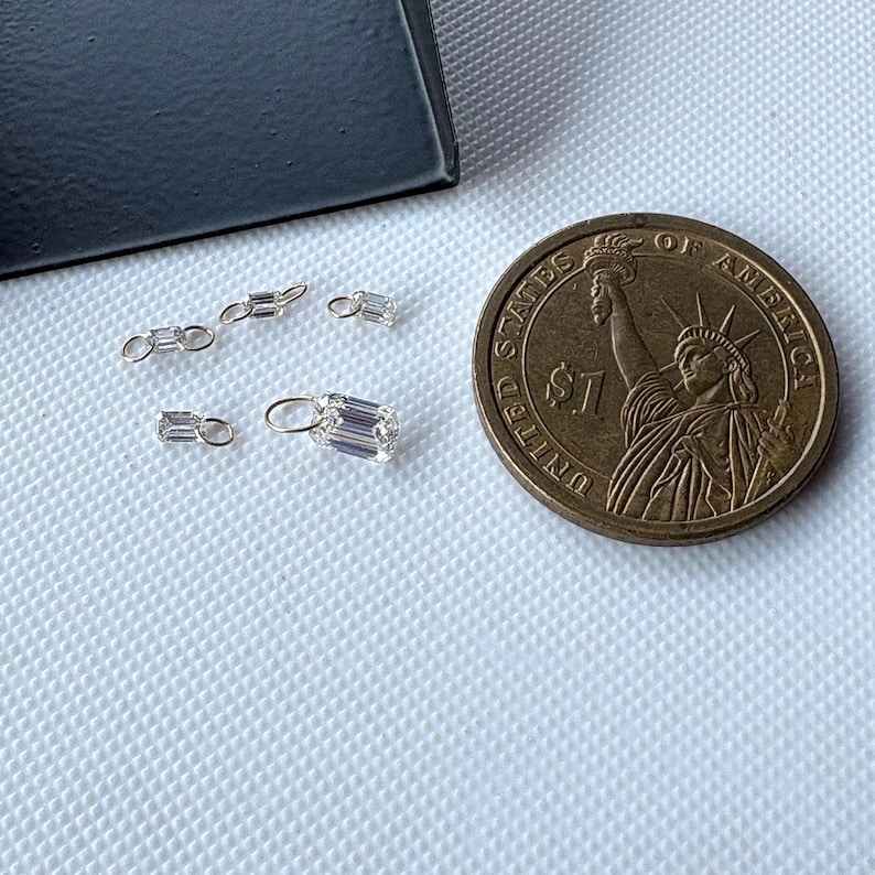 May include: A collection of small, clear, rectangular-cut gemstones with silver-colored settings. The gemstones are scattered on a textured white surface next to a US one-dollar coin. The gemstones vary in size and shape, some with loops for hanging.