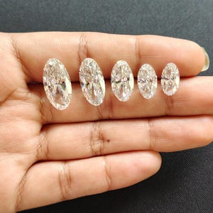 May include: Five clear, colorless, oval-cut gemstones arranged in a row by size from largest to smallest. Each stone features multiple facets that reflect light, creating a brilliant, sparkling appearance with a transparent, crystalline structure.