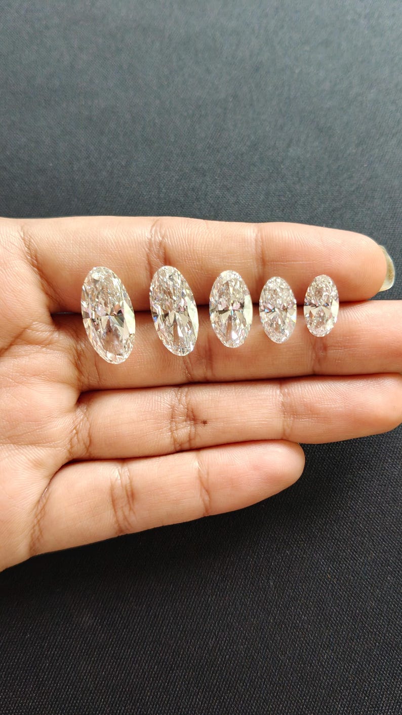 May include: Five clear, colorless, oval-cut gemstones arranged in a row by size from largest to smallest. Each stone features multiple facets that reflect light, creating a brilliant, sparkling appearance with a transparent, crystalline structure.