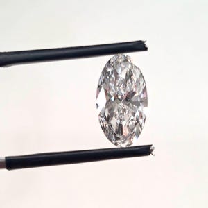 May include: A marquise-cut loose diamond with a clear, colorless appearance and multiple facets that reflect light. The gemstone is held vertically between two thin, black metal prongs, showcasing its elongated, pointed oval shape and brilliance.