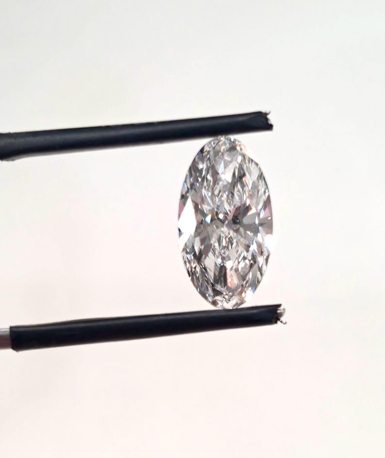 May include: A marquise-cut loose diamond with a clear, colorless appearance and multiple facets that reflect light. The gemstone is held vertically between two thin, black metal prongs, showcasing its elongated, pointed oval shape and brilliance.