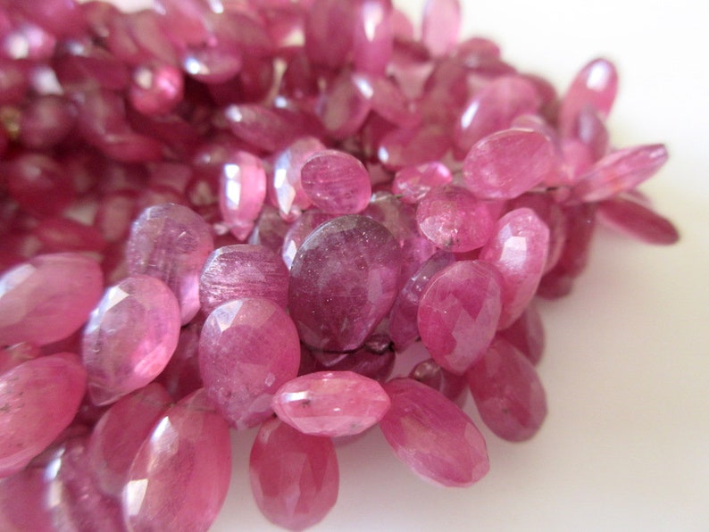 May include: A strand of pink faceted gemstone beads. The beads are oval shaped and have a smooth, polished finish.
