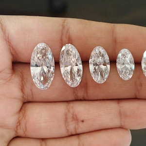 May include: Five clear, colorless, oval-cut gemstones arranged in a row by size from largest to smallest. Each stone features multiple facets that reflect light, creating a brilliant, sparkling appearance with a transparent, crystalline texture.