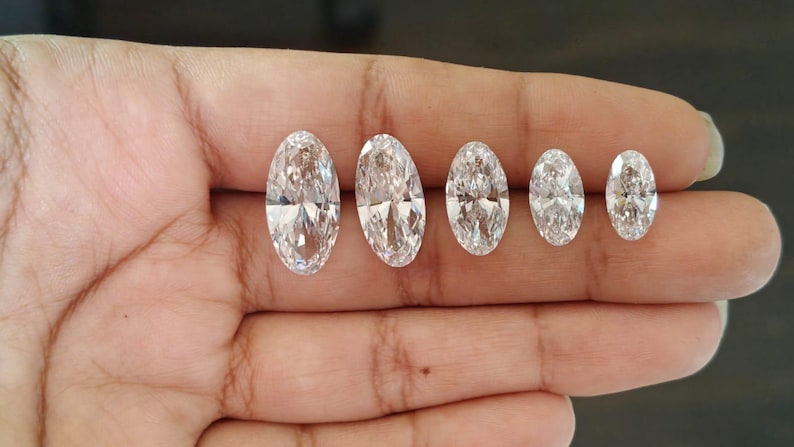 May include: Five clear, colorless, oval-cut gemstones arranged in a row by size from largest to smallest. Each stone features multiple facets that reflect light, creating a brilliant, sparkling appearance with a transparent, crystalline texture.