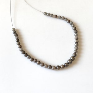 May include: Strung beads in varying sizes, featuring a metallic gray color. The beads are on a thin, silver-colored string, arranged in a curved shape against a white background. The beads appear to be made of a polished stone.