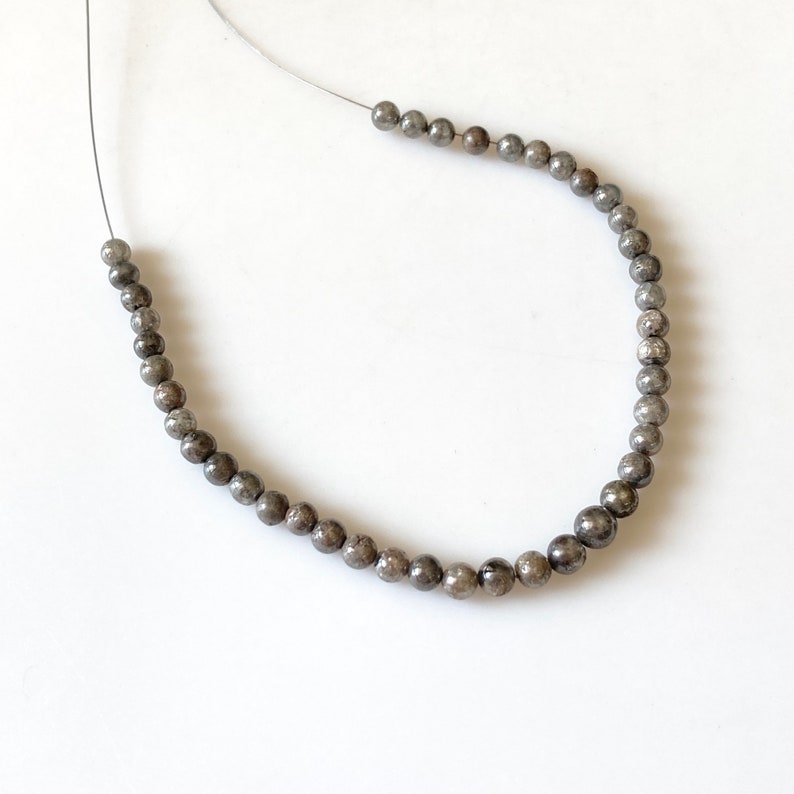 May include: Strung beads in varying sizes, featuring a metallic gray color. The beads are on a thin, silver-colored string, arranged in a curved shape against a white background. The beads appear to be made of a polished stone.