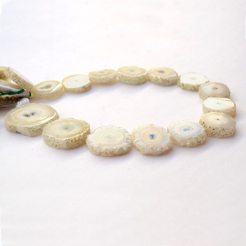 May include: A strand of white and cream colored agate beads, each bead is a slice of a geode with a crystal pattern. The beads are strung on a white cord and are arranged in a circle.