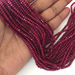 Puede incluir: Una hebra de cuentas de rubí rojo intenso, facetadas y pulidas, enhebradas en un fondo blanco. Las cuentas son pequeñas y de tamaño uniforme, creando un aspecto delicado y elegante.