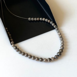 May include: A strand of small, round, gray beads, possibly gemstones, strung on a thin, silver-colored wire. The beads have a speckled appearance and are displayed against a black tray and a white surface.