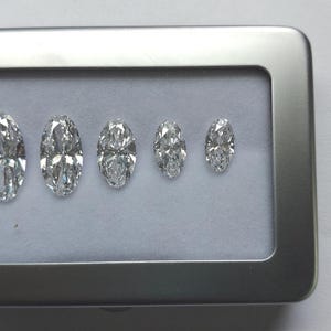 May include: Five clear, faceted, marquise-cut gemstones arranged in a row by size, from largest to smallest, inside a rectangular silver-colored metal display case with a white interior lining. The stones exhibit high clarity and brilliance.