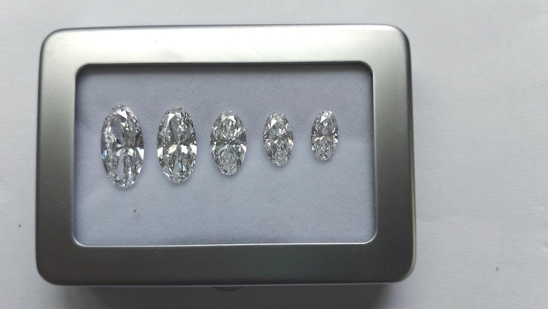 May include: Five clear, faceted, marquise-cut gemstones arranged in a row by size, from largest to smallest, inside a rectangular silver-colored metal display case with a white interior lining. The stones exhibit high clarity and brilliance.