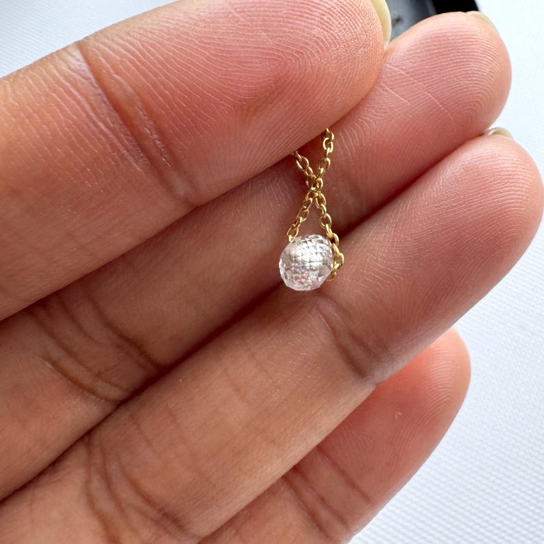 K&ouml;nnte beinhalten: Eine zarte goldene Halskette mit einem einzelnen, kleinen, klaren, facettierten und runden Edelsteinanh&auml;nger. Die Kette bildet eine V-Form, w&auml;hrend sie mit der Oberseite des funkelnden, durchscheinenden Steins verbunden ist, was ein schlichtes Schmuckst&uuml;ck ergibt.