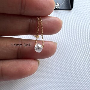 K&ouml;nnte beinhalten: Eine klare, facettierte, runde Edelsteinperle, die an einer zarten, goldfarbenen Metallkette h&auml;ngt. Die Perle weist ein 1,5 mm gro&szlig;es Bohrloch auf. Das Schmuckelement wird im Detail gezeigt und hebt die reflektierende Oberfl&auml;che des Steins hervor.