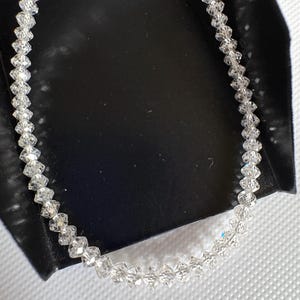 Puede incluir: Un delicado collar de cristal transparente. El collar est&aacute; compuesto por numerosos cristales peque&ntilde;os y facetados ensartados. Los cristales est&aacute;n dispuestos en un patr&oacute;n lineal, creando un accesorio brillante y elegante. El collar se muestra sobre una superficie negra.