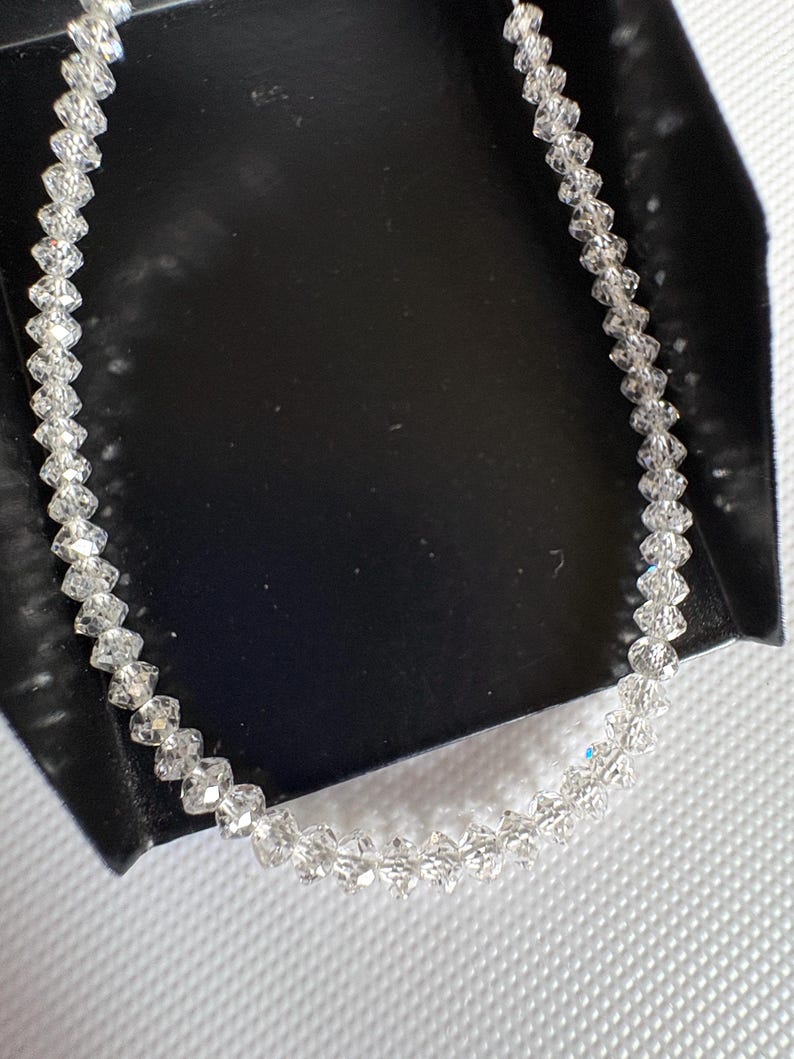 Puede incluir: Un delicado collar de cristal transparente. El collar est&aacute; compuesto por numerosos cristales peque&ntilde;os y facetados ensartados. Los cristales est&aacute;n dispuestos en un patr&oacute;n lineal, creando un accesorio brillante y elegante. El collar se muestra sobre una superficie negra.