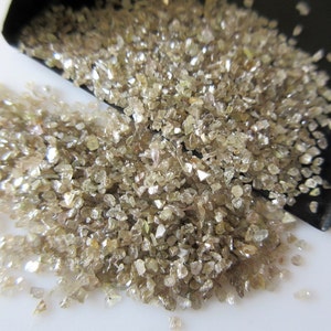 May include: A pile of small, rough, light brown diamonds. The diamonds are scattered on a white surface and are illuminated by a bright light.