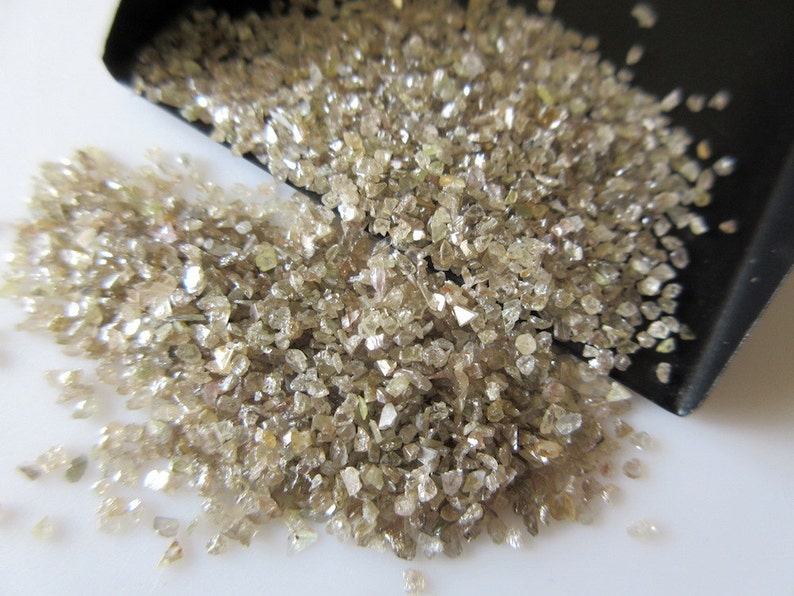 May include: A pile of small, rough, light brown diamonds. The diamonds are scattered on a white surface and are illuminated by a bright light.