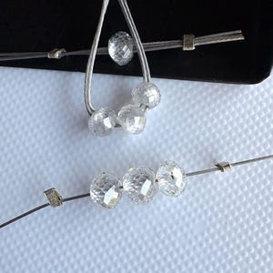 K&ouml;nnte beinhalten: Klare, facettierte runde Perlen auf d&uuml;nnem, silberfarbenem Metalldraht. Die Perlen sind in einer Schlaufe und einer geraden Linie angeordnet und durch kleine metallische Quetschperlen getrennt. Die transparenten, facettierten Oberfl&auml;chen reflektieren das Licht.