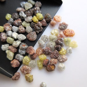 May include: A collection of rough, uncut diamonds in various shades of brown, gray, and yellow. The diamonds are scattered on a white surface.