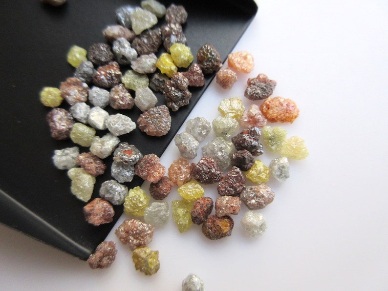 May include: A collection of rough, uncut diamonds in various shades of brown, gray, and yellow. The diamonds are scattered on a white surface.