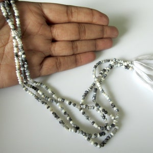 Op de afbeelding: Een meerstrengs ketting bestaande uit kleine, facetgeslepen kralen in wit, grijs en zwart. De ketting heeft een kwast van witte draden. De kralen zijn geregen, waardoor een delicaat en elegant accessoire ontstaat, geschikt voor diverse outfits.