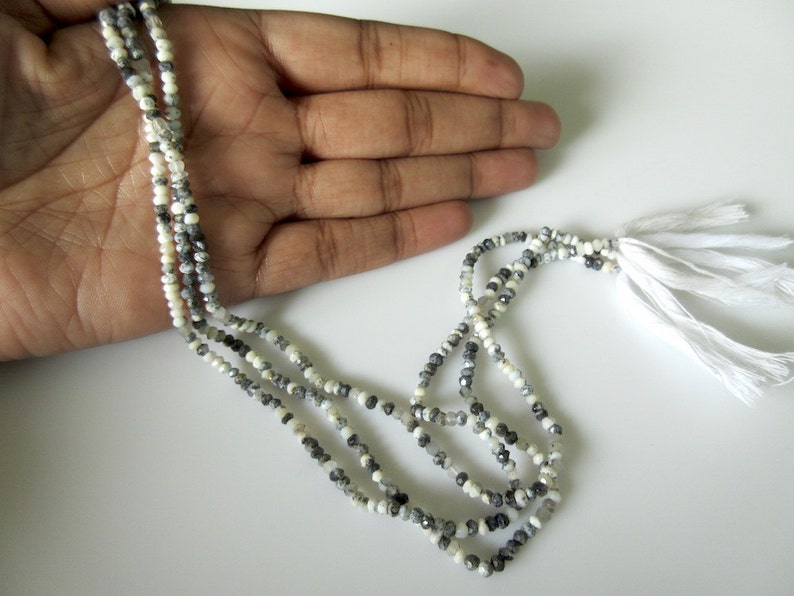 Op de afbeelding: Een meerstrengs ketting bestaande uit kleine, facetgeslepen kralen in wit, grijs en zwart. De ketting heeft een kwast van witte draden. De kralen zijn geregen, waardoor een delicaat en elegant accessoire ontstaat, geschikt voor diverse outfits.