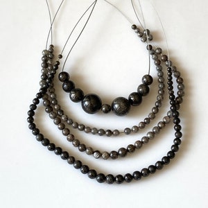 May include: Four strands of beaded necklaces in varying sizes and shades of gray and black. The beads range from small to large, with the largest beads in the center of the top strand. The necklaces are strung on thin, dark cords.