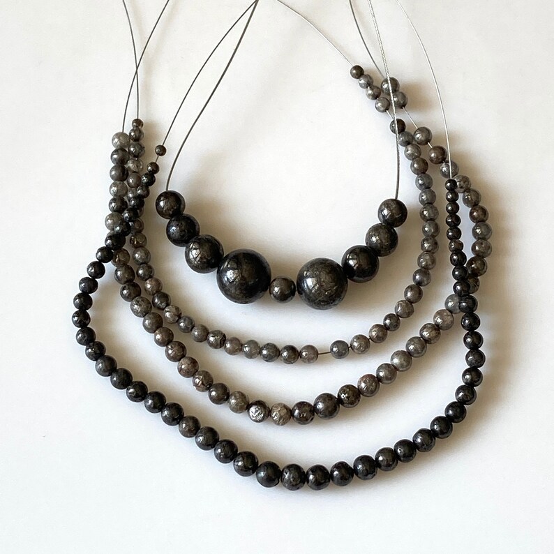 May include: Four strands of beaded necklaces in varying sizes and shades of gray and black. The beads range from small to large, with the largest beads in the center of the top strand. The necklaces are strung on thin, dark cords.