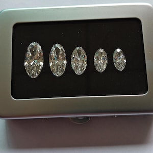 May include: A rectangular silver-colored metal display case with a hinged lid and a black interior insert. Five clear, oval-cut faceted gemstones are arranged in a row by size, from largest to smallest, resting securely within the black foam base.