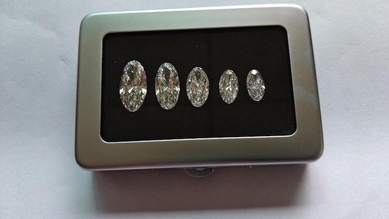 May include: A rectangular silver-colored metal display case with a hinged lid and a black interior insert. Five clear, oval-cut faceted gemstones are arranged in a row by size, from largest to smallest, resting securely within the black foam base.