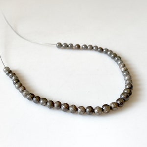 May include: Strung beads in a dark gray color, possibly pyrite, are displayed against a white background. The beads are small, round, and evenly spaced on a thin, clear string, suggesting jewelry making.