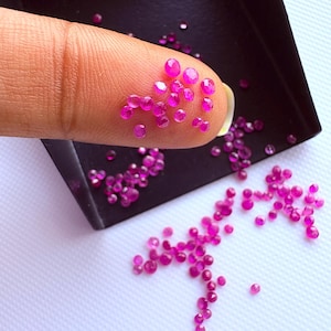 May include: A collection of small, round, faceted, vibrant pinkish-red gemstones. Several of these loose stones are resting on a fingertip, while others are scattered across a black tray and a white surface, showcasing their reflective surfaces.