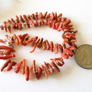 May include: A strand of coral beads with a reddish-orange color. The beads are irregular in shape and size, and some have a rough texture. A US quarter is included for scale.