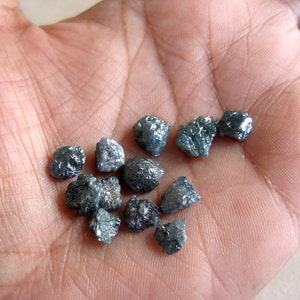 Könnte beinhalten: Eine Sammlung kleiner, dunkelgrauer, grob geschliffener Diamanten mit einem bläulich-grünen Farbton. Die Diamanten sind auf einer Hand verstreut.
