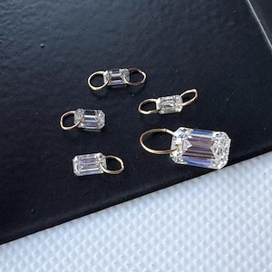 May include: Five small, rectangular, clear crystal charms with gold-colored loops. The crystals have a faceted cut, catching the light. The charms are arranged on a dark surface, creating a contrast that highlights their sparkle. These charms are suitable for jewelry making.