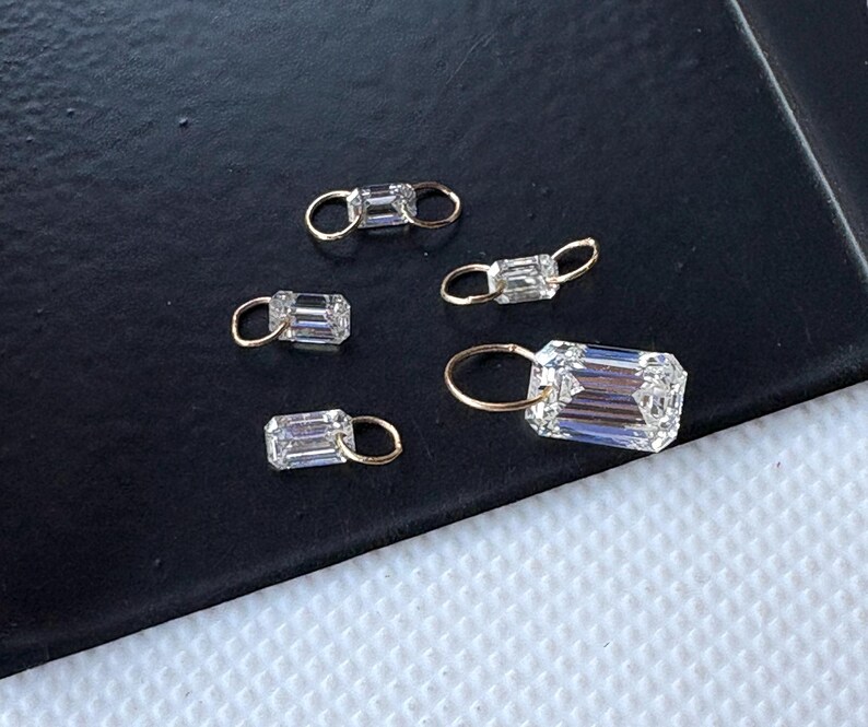 May include: Five small, rectangular, clear crystal charms with gold-colored loops. The crystals have a faceted cut, catching the light. The charms are arranged on a dark surface, creating a contrast that highlights their sparkle. These charms are suitable for jewelry making.