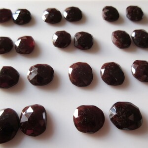 May include: A collection of faceted, dark red gemstones, possibly garnets, arranged on a white surface. The stones have a polished, reflective surface and a deep, rich color. The shapes vary slightly, with some appearing more rounded and others more elongated.