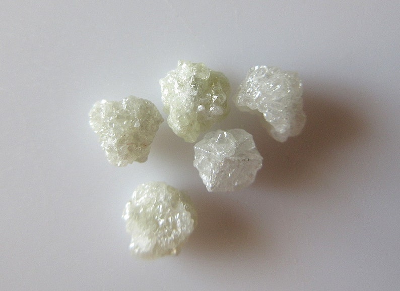 Puede incluir: Cinco diamantes en bruto, blancos y transl&uacute;cidos. Los diamantes est&aacute;n agrupados en una superficie blanca.