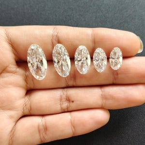 May include: Five clear, colorless, oval-cut gemstones arranged in a row by size from largest to smallest. Each stone features a faceted surface that reflects light, creating a brilliant, sparkling appearance with sharp, defined edges.