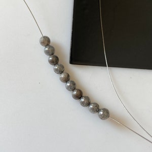 May include: A strand of gray, spherical beads on a thin, silver wire. The beads have a textured surface and are arranged in a line. A black, rectangular object is in the background.