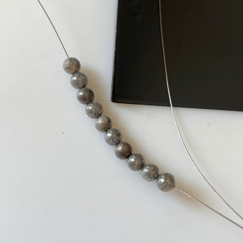 May include: A strand of gray, spherical beads on a thin, silver wire. The beads have a textured surface and are arranged in a line. A black, rectangular object is in the background.