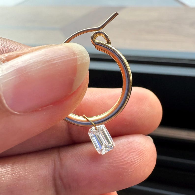May include: A gold hoop earring with a small, rectangular, clear gemstone pendant. The earring is held in a person's hand, showcasing the delicate design and the sparkle of the gemstone. The hoop is approximately 1 inch in diameter.