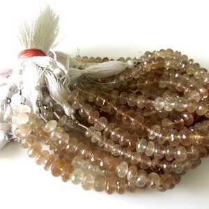 May include: A strand of light brown faceted gemstone beads. The beads are oval shaped and have a translucent appearance. The beads are strung on a white thread and have a tassel at the end.