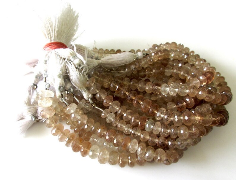 May include: A strand of light brown faceted gemstone beads. The beads are oval shaped and have a translucent appearance. The beads are strung on a white thread and have a tassel at the end.