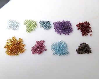 May include: Assortment of small, round, faceted gemstones in various colors, including blue, green, purple, red, brown, and orange. The gemstones are arranged in rows on a white surface.