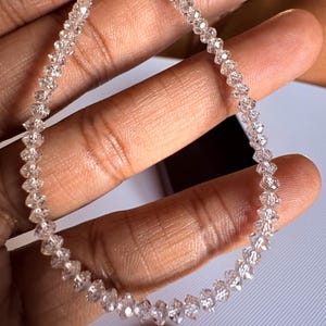 Puede incluir: Un hilo de cuentas claras y facetadas, posiblemente piedras preciosas, dispuestas en forma circular. Las cuentas son peque&ntilde;as y brillantes, con un corte uniforme. La imagen est&aacute; bien iluminada, resaltando la claridad y el brillo de las cuentas. Una moneda es visible en el fondo.