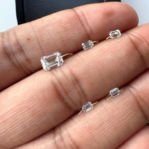 May include: A collection of small, clear, rectangular-cut diamond earrings with gold-colored posts. The earrings are displayed against a neutral background, showcasing their sparkle and design. The diamonds are set in a simple, elegant style.