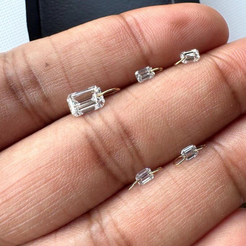 May include: A collection of small, clear, rectangular-cut diamond earrings with gold-colored posts. The earrings are displayed against a neutral background, showcasing their sparkle and design. The diamonds are set in a simple, elegant style.