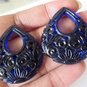 May include: Two dark blue, teardrop-shaped pendants with detailed floral carvings. Each pendant features a central teardrop-shaped opening. The carvings create a textured surface. These pendants are likely for jewellery making.